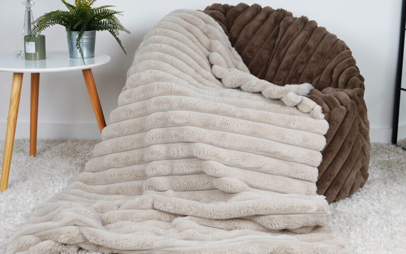 The image shows a faux fur throw blanket draped over a faux fur bean bag chair. The bean bag chair is a light brown colour, and the throw blanket is a light beige colour. The throw blanket has a ribbed texture and is very soft. The bean bag chair is placed in a living room setting, with a white coffee table and a potted plant nearby.