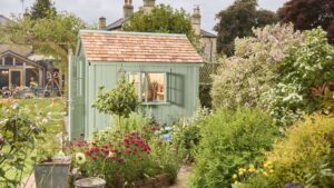 The primary focus is the potting shed, painted in a soft, muted green. It has a traditional design with vertical plank siding and a pitched roof covered in small, rectangular cedar shingles. The double doors have simple panelled detailing.