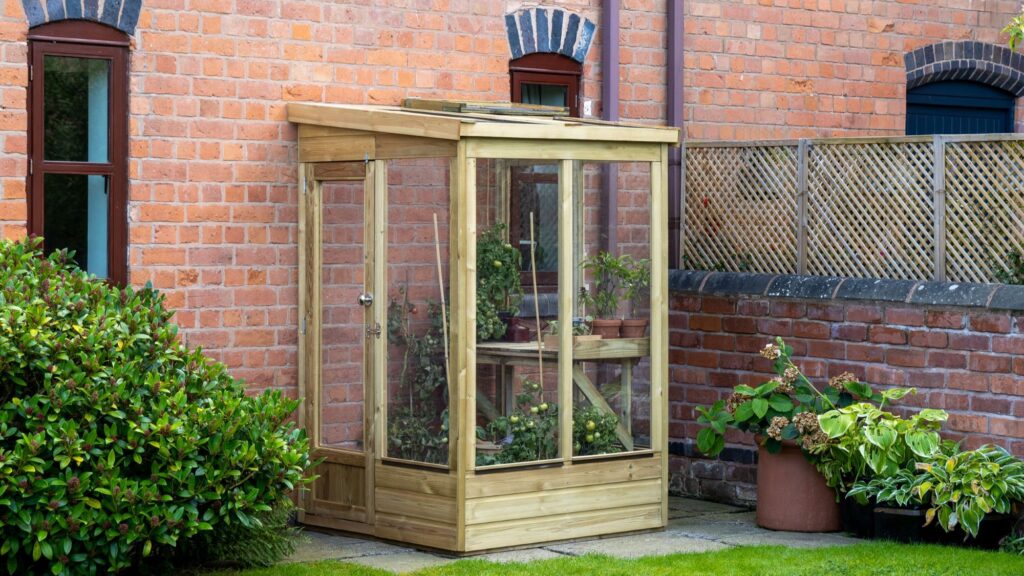 Outdoor image of a wooden lean-to greenhouse with a single door and clear panels, positioned against a brick wall with arched windows. Potted plants are visible inside.