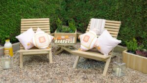 Two slatted wooden chairs and a small slatted wooden table arranged on a gravel surface. The chairs have cushions on them, and there are a few decorative items on the table, along with lanterns on the ground nearby.