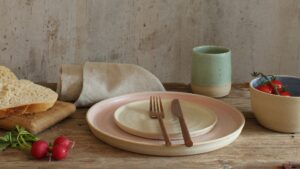 A stack of two ceramic plates. The larger plate has a light beige exterior and a soft pink interior. The smaller plate is a creamy white. A fork and knife rest on the smaller plate.