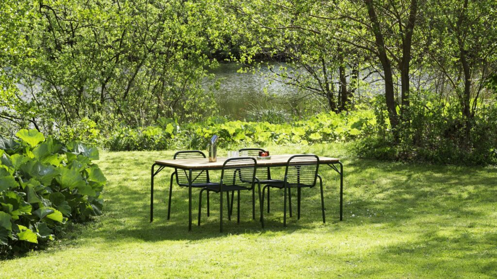 A vibrant outdoor scene features a wooden rectangular dining table with four black metal chairs, set on a lush green lawn.