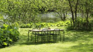A vibrant outdoor scene features a wooden rectangular dining table with four black metal chairs, set on a lush green lawn.