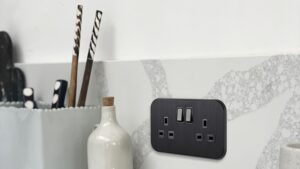 A sleek, black double UK plug socket (13A) is mounted on a grey and white marble-effect splashback in a modern kitchen.