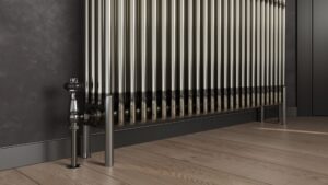 A bare metal, multi-column radiator stands on a light wooden floor with dark grey skirting boards and a dark grey wall behind it. To the left, a silver valve and pipe connect to the radiator, and on the right, two matching silver feet support it.
