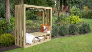 A natural wood-stained pergola with an integrated bench seat is positioned on a vibrant green lawn. The pergola has a rectangular shape with slatted sides and an open roof, from which hangs a string of decorative globe lights. On the bench, there are several cushions in warm tones of orange, brown, and cream.