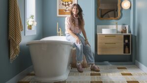 A woman with long, dark hair is seated on the edge of a white, freestanding bathtub in a bathroom. She is wearing a light-coloured, long-sleeved top and blue jeans. To her right, there is a wooden wall-mounted vanity unit with a white circular sink and a gold-coloured faucet.
