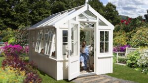 A charming, bright white greenhouse stands amidst a vibrant, sunlit garden. The greenhouse features a classic design with a gabled roof, a significant portion of which, along with the upper sections of its walls, is made of glass, allowing ample light to penetrate. The lower part of the greenhouse has solid, painted walls.