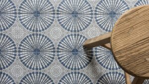 A bird’s eye view of a patterned floor tile. The light grey and blue tile has a circular geometric pattern. A small wooden stool is also seen.